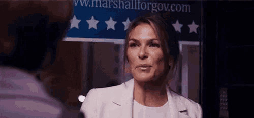 a woman in a white suit is talking to a man in front of a sign that says ' shallforgov ' .