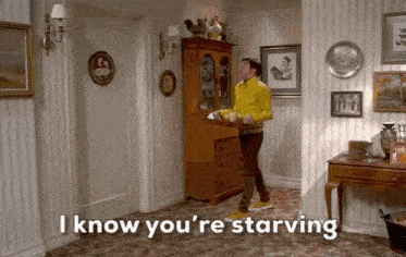 a man in a yellow jacket is holding a tray in a living room and says i know you 're starving