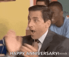 a man in a suit and tie is sitting at a table with his hands in the air and saying happy anniversary .
