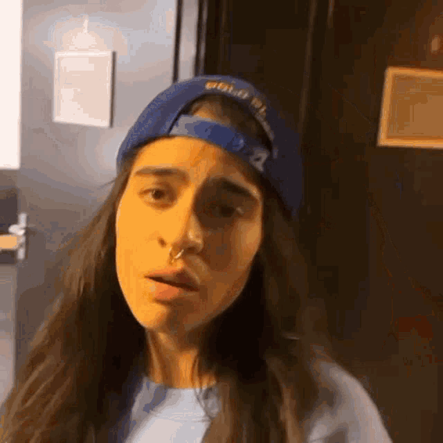 a woman with long hair and a nose ring is wearing a blue hat and making a funny face .