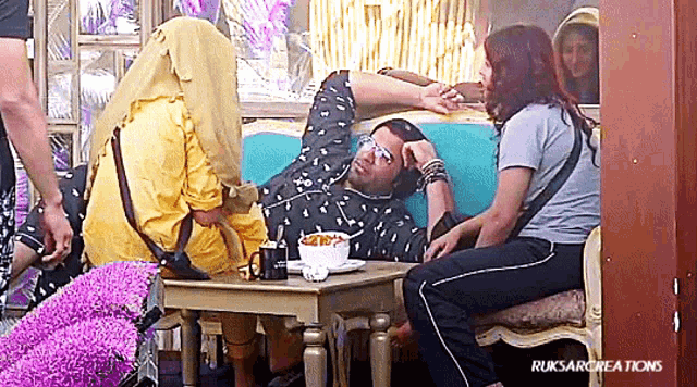 a group of people are sitting around a table with a man laying on a couch