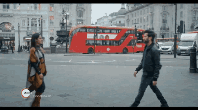 a man and a woman are walking in front of a red double decker bus with a zeus logo on the bottom