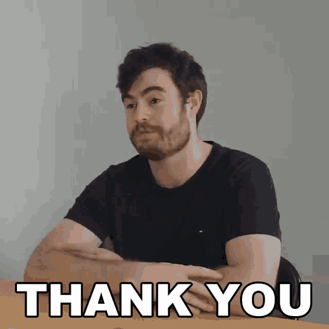 a man with a beard is sitting at a table with his arms crossed and the words thank you above him