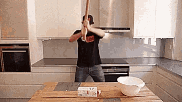 a man in a black shirt is holding a wooden bat in a kitchen .