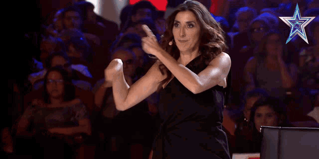 a woman in a black dress is dancing in front of a crowd with a blue star in the corner