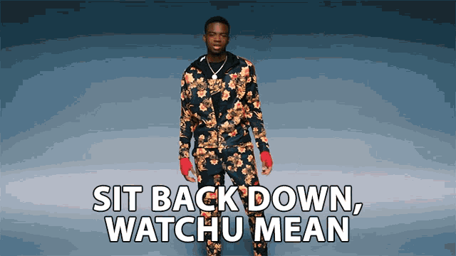 a man in a floral suit stands in front of a blue background with the words sit back down watchu mean