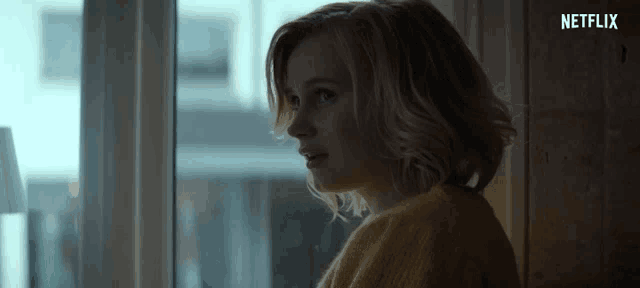 a woman in a yellow sweater is looking out a window with a netflix logo in the background