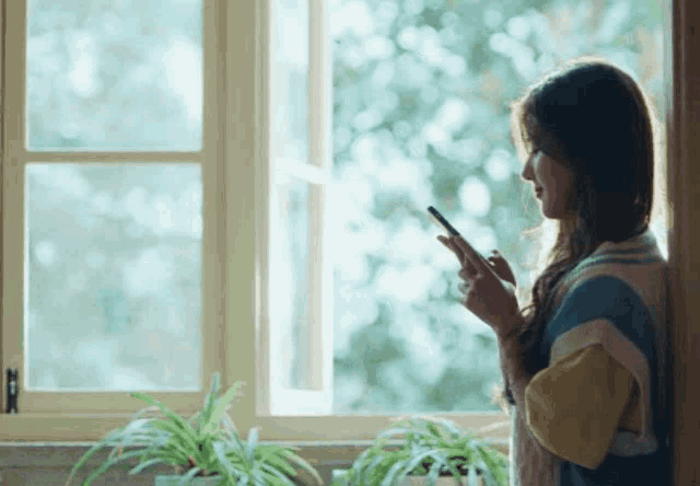 a woman leaning against a wall looking out a window while holding a cell phone
