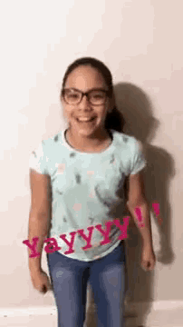 a little girl wearing glasses and a blue shirt is standing in front of a white wall .