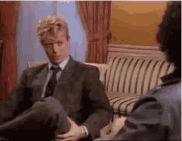 a man in a suit and tie is sitting on a couch with his legs crossed and talking to another man .