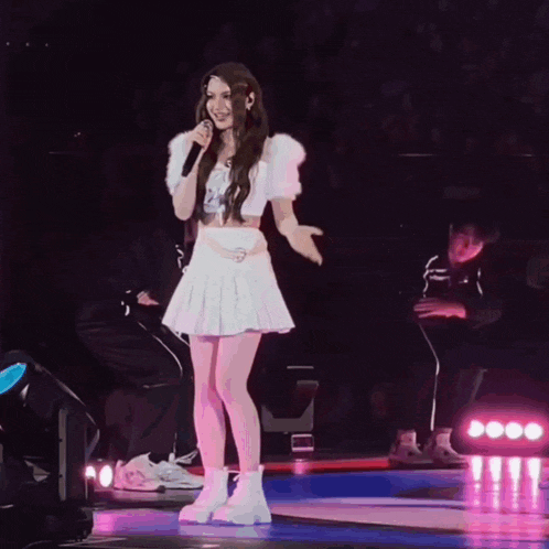 a woman in a white pleated skirt is singing into a microphone on a stage