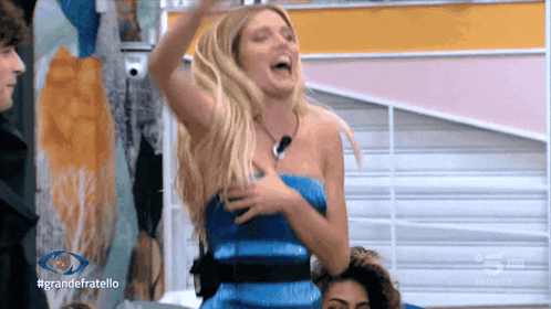 a woman in a blue dress is laughing in front of a sign that says #grandefratello