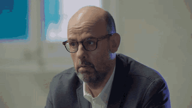 a bald man wearing glasses and a suit looks at something
