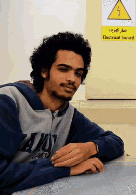 a man leaning on a table in front of a yellow sign that says electrical hazard