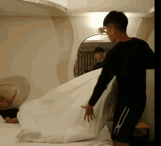 a man in a black shirt is standing next to a bed with a white comforter