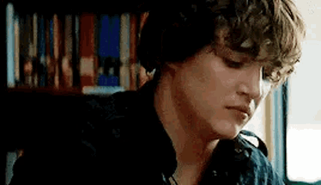 a young man with curly hair is sitting in front of a bookshelf in a library .