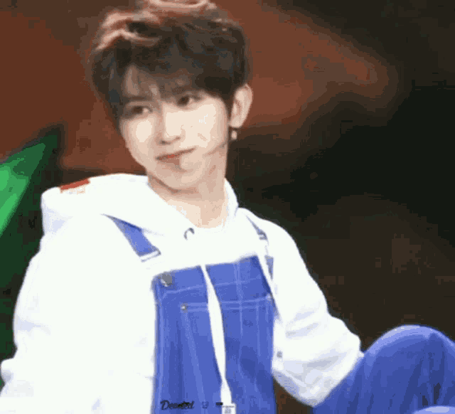 a young man wearing blue overalls and a white hoodie is sitting on a stage .