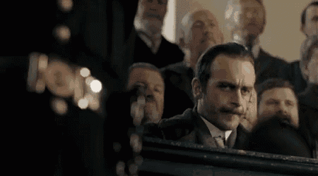 a man with a mustache is sitting in front of a crowd of people in a dark room .