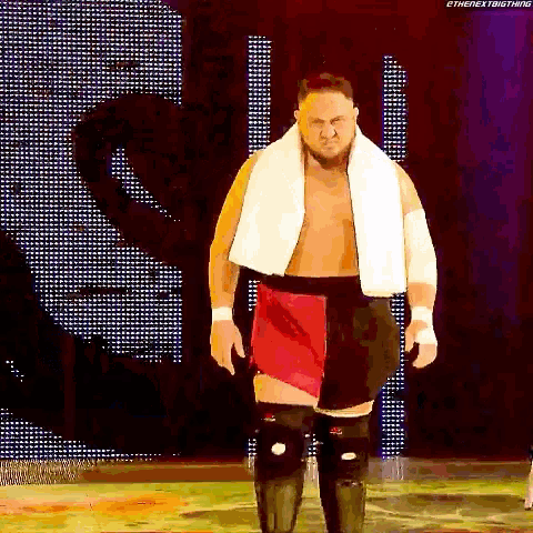 a wrestler with a white towel around his neck is walking on a stage .