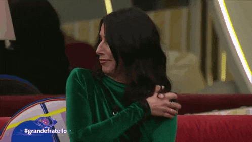 a woman in a green top is sitting on a red couch with #grandefratello written on the screen behind her