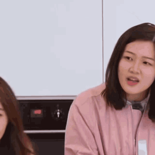 a woman in a pink jacket is sitting in a kitchen with other women .
