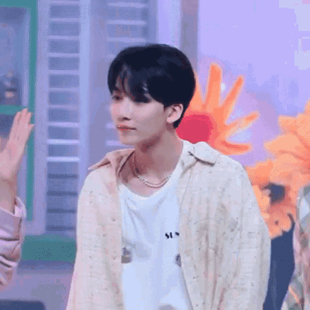 a young man wearing a white shirt and a pearl necklace is standing in front of a bunch of flowers .