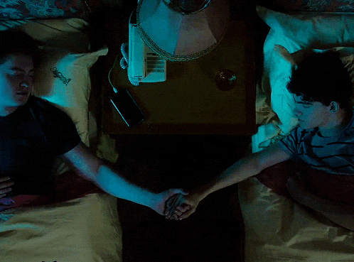 two men laying on a bed holding hands with a lamp on the nightstand behind them