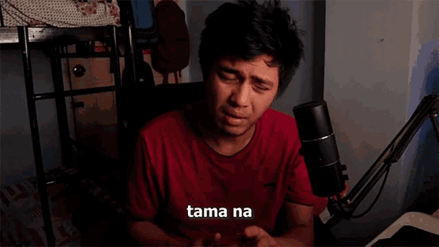 a man in a red shirt is sitting in front of a microphone and the word tama na is on the screen
