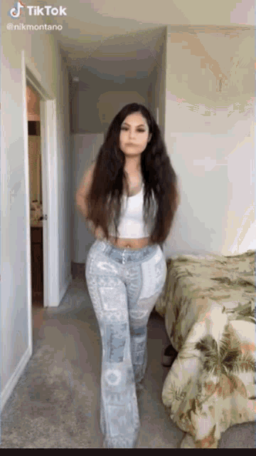 a woman in a white tank top and blue pants is standing in a room with a bed .