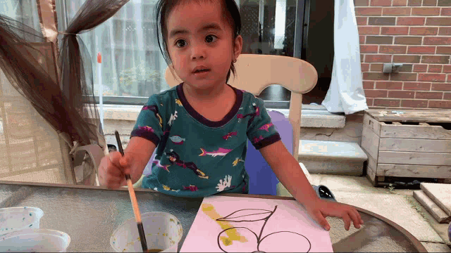 a little girl in a blue shirt with sharks on it paints a picture with a brush