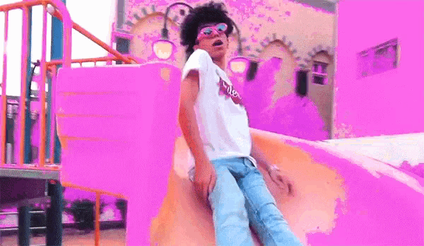 a person wearing sunglasses is riding a pink slide at a playground
