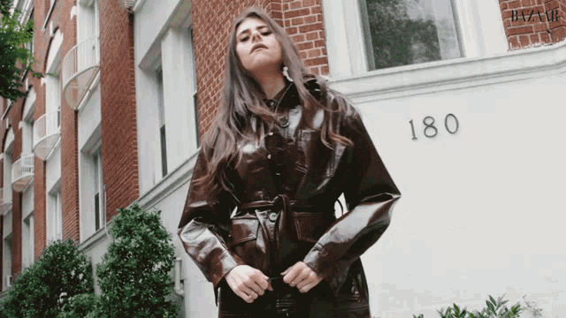 a woman in a brown leather jacket stands in front of a building that says 180