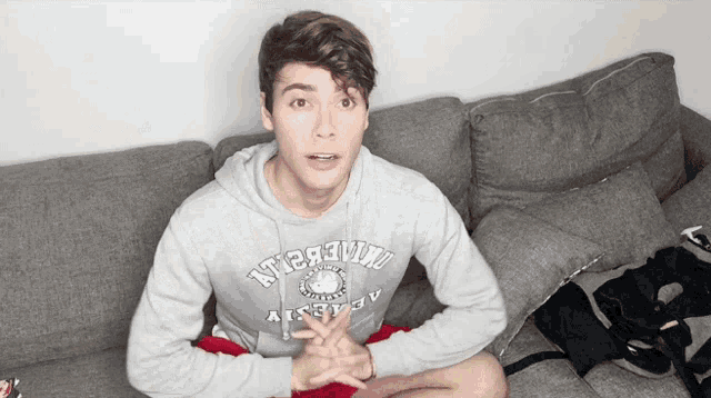 a young man is sitting on a couch wearing a grey hoodie that says ' university ' on it