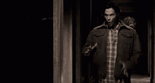 a man in a plaid shirt and jacket is standing in a dark hallway .