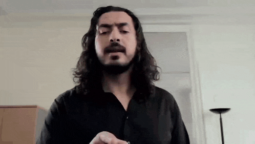 a man with long hair and a beard is standing in front of a window and looking at the camera .