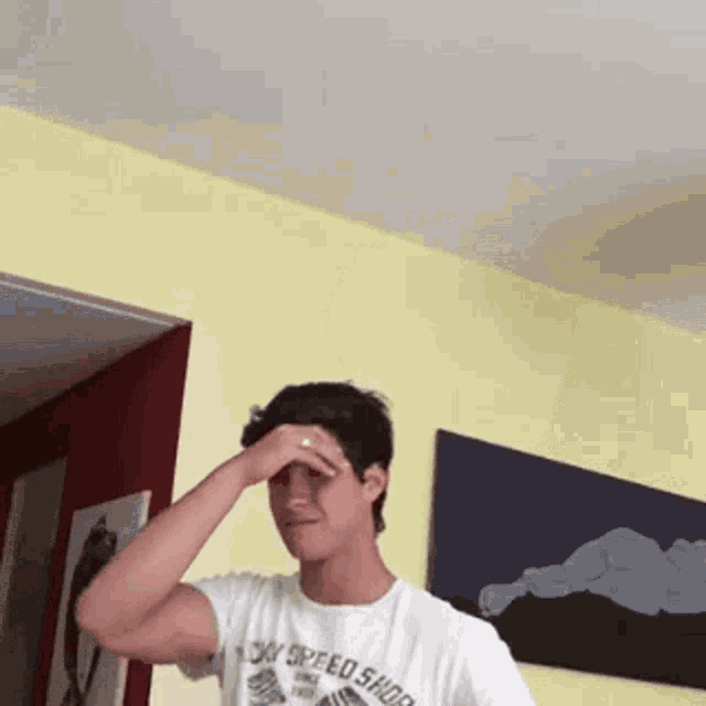 a young man wearing a white t-shirt that says on speed shop is covering his eyes with his hand .