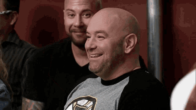 a bald man with a beard is smiling while sitting next to another man .