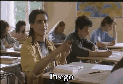a man with a mullet is sitting at a desk in a classroom with other students and says prego .