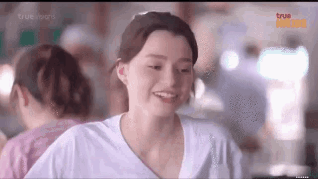 a woman in a white t-shirt is smiling and looking at the camera in a crowded room .