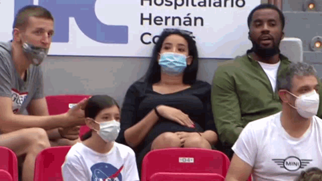 a group of people wearing face masks sit in a stadium with a sign that says hospitalario hernan