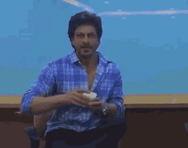 a man in a blue plaid shirt is sitting at a table holding a cup .