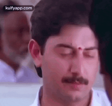 a man with a mustache and a red dot on his forehead is making a funny face with his eyes closed .