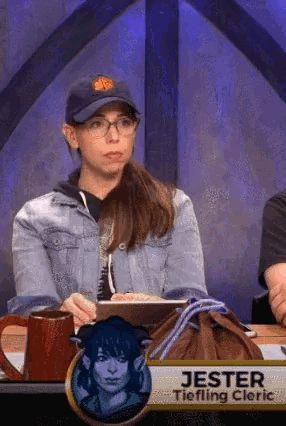 a woman wearing a hat and glasses is sitting at a table with a sign that says jester tiefling cleric