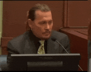a man in a suit and tie is sitting at a podium in front of a microphone with the word no written on it .