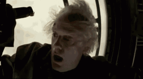 a man with white hair is looking out of a window with his mouth open and his hair blowing in the wind .