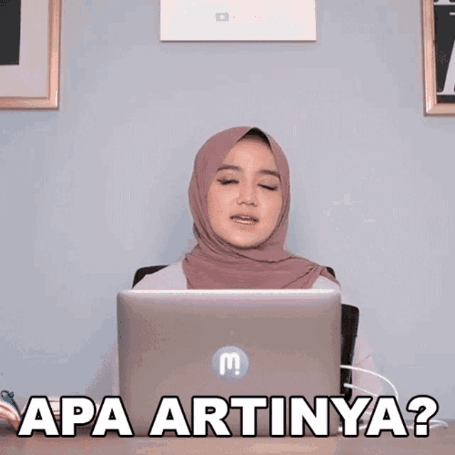 a woman wearing a hijab sits in front of a laptop and says apa artinya