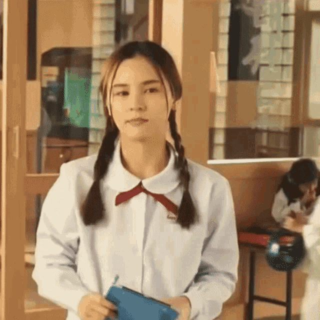a girl in a school uniform with pigtails is standing in a classroom holding a purse .