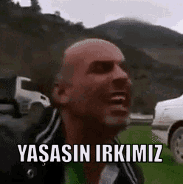 a bald man is screaming in front of a mountain with the words yasasin irkimiz written on his face .