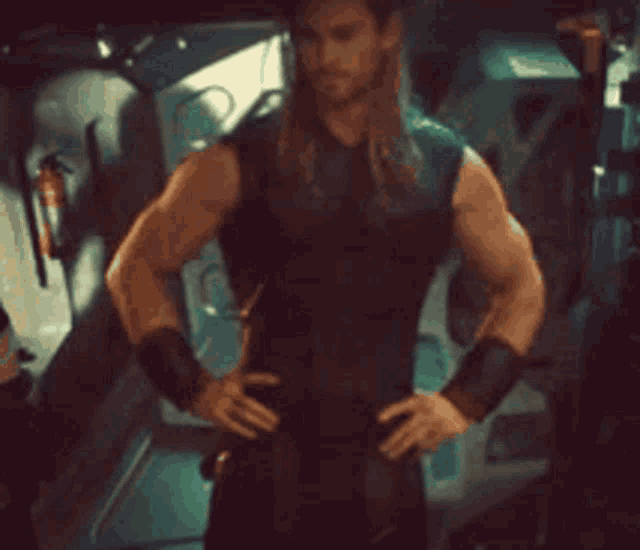 a man with long hair and a beard is standing with his hands on his hips in a dark room .
