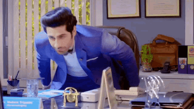 a man in a blue suit is sitting at a desk with a name plate that says vedanta tripathi
